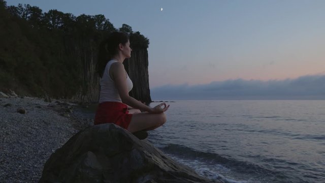 Woman at sunset meditates in lotus position sitting on a large stone near the sea.