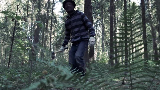 Serial maniac with axe searching for victim in forest. Bloody killer, murderer, mad man in scary   mask and hat. Halloween costume, horror concept.