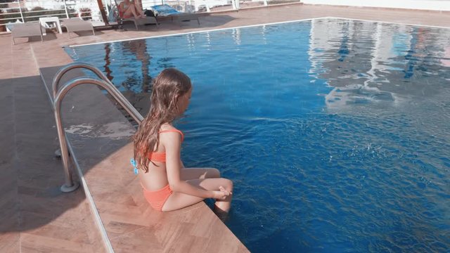 Happy child in swimwear playing in pool. High angle view of cute preteen girl in swimsuit sitting with feet in swimming pool and playing with water at summertime