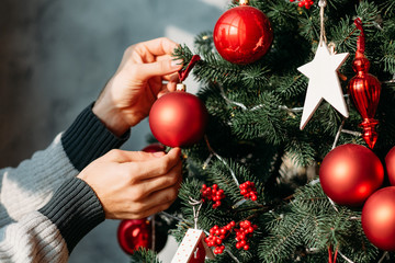 Winter holidays concept. Closeup of man hands decorating green fir tree with red ball ornaments.