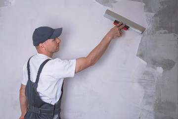 Worker in overalls plastering a wall with finishing putty using a putty knife. Repair work and...