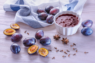 A white ceramic bowl with homemade plum jam, fresh ripe plums, crumpled towel, badyan seeds and dry carnation grains on a wooden table in the morning light