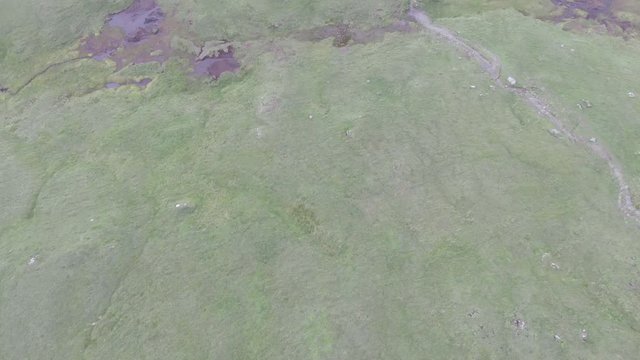 An aerial forward reveal footage of a grassy mountain plateau rocky summit with small pond and mountain range in the background under a cloudy sky
