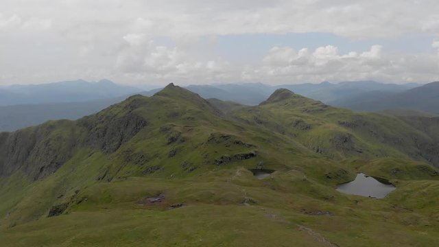 An aerial forward footage of a grassy mountain plateau rocky summit with small pond and mountain range in the background under a cloudy sky