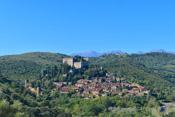 Fototapeta na wymiar French quaint town at the foot of a castle on a hill surrounded by vineyards and greenery. Gorgeous Pyrenees mountains at the background. Castelnou, south of France