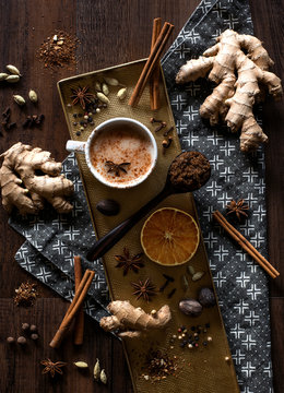 chai tea spices flat lay, chai tea latte with ginger root, cinnamon sticks, cardamom pods, star anise, whole nutmeg, peppercorns, whole cloves on a golden tray and grey patterned cloth napkin on table