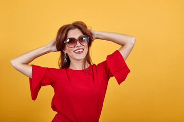 Happy girl wearing sunglasses smiling, keeping her hands behind your head. Gorgeous woman standing over yellow wall with glasses and cheerful