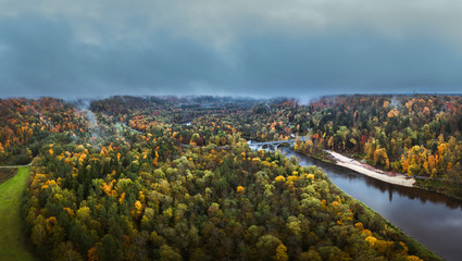 Fototapeta na wymiar Valley of colorful trees covered fog at early mourning. Rainy weather with storm clouds in the sky. Picturesque panorama with river Gauja curving through the valley. 