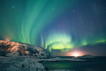 Weitwinkelaufnahme von Aurora und dem Sternenhimmel mit schneebedeckten Hügeln und dem See