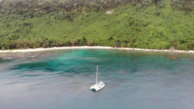Aerial view of a catamaran yacht anchored in clear blue water and white sand off a tropical island with coral reef and palm trees in Vanuatu