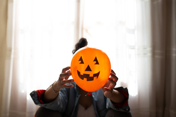 Witch holding a pumpkin balloon with her hands and covering her face. Young woman celebrating halloween. Helium party balloons.