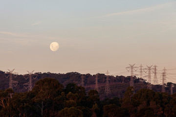 Full moon setting during twilight over natural landscape below with electricity pylons.
