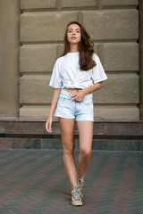 Full-length portrait of trendy stylish girl in the city. She walks on the street and turn around on camera. Wearing in white t-shirt short denim shorts and sneakers. Truly emotions. Place for text.