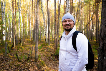Naklejka na ściany i meble Hiker with a backpack looks at the camera and admires the autumn forest. Autumn nature scenery. Portrait of an adult male back standing outdoor.