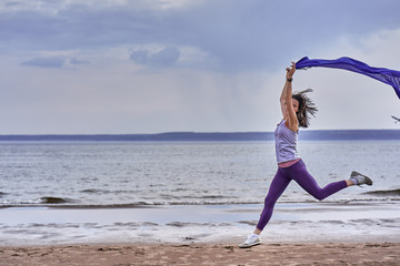 Obraz premium Young slim brunette woman jumping while jogging while holding a blue scarf in her hands. A woman is engaged in gymnastics in the spring morning on the sandy bank of a large river. Cloudy morning.