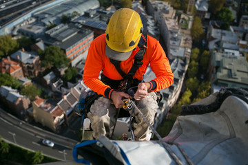 Fototapeta premium Close up pic of male rope access jobs worker wearing yellow hard hat, long sleeve shirt, safety harness, working, at height abseiling down from the high rise building site, Sydney city, Australia