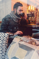 young guy with a beard in a sweater works at a computer in a cozy warm cafe with a cup of coffee