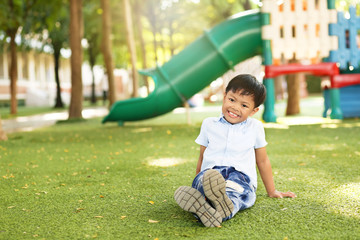 An Asian young boy sitting on green artificial grass with smiling at a playground.