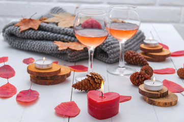 Small burning candles, two glasses with rose wine, cones, dry red leaves, a gray scarf knitted on a white wooden table. Hello, Autumn. Cozy autumn background.