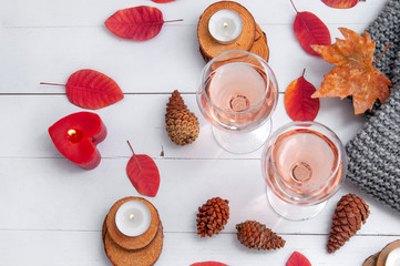 Small burning candles, two glasses with rose wine, cones, dry red leaves, a gray scarf knitted on a white wooden table. Hello, Autumn. Cozy autumn background.