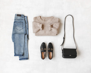 Blue jeans, beige knitted oversize sweater, small cross body bag, black loafers or flat shoes on grey background. Overhead view of women's casual day outfit. Flat lay, top view. Women clothes.