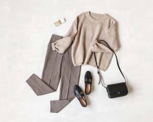 Brown pants in check, beige knitted oversize sweater, cross body bag, black loafers or flat shoes on grey background. Overhead view of women's casual day outfit. Flat lay, top view. Women clothes.