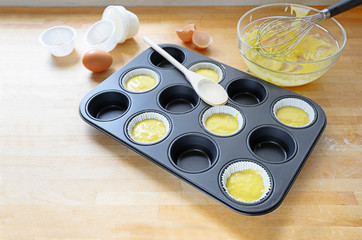 muffin tin, partly filled with paper cups and dough for cupcake baking on a wooden kitchen table, copy space