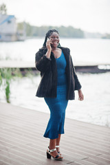 African american dark skinned plus size model posed in a blue shiny dress and black fur coat against lake and speaking by mobile phone.