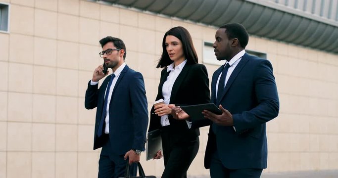 African American businessman and Caucasian businesswoman walking the street and discussing business and work while their Arabian male colleague walking by and talking on the phone. - Powered by Adobe
