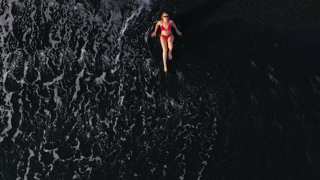 Top view of a woman in a red swimsuit lying on a black beach on the surf line. Coast of the island of Tenerife, Canary Islands, Spain.