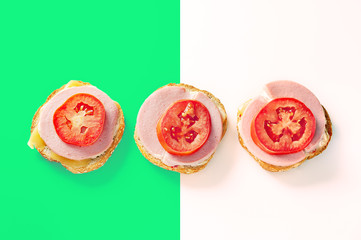 Three Open Sandwiches Made of One Slice of Bread with Sesame Seeds in Crust, and Slices of Cheese, Cooked Sausage and Tomato on Top. Green and White Background.