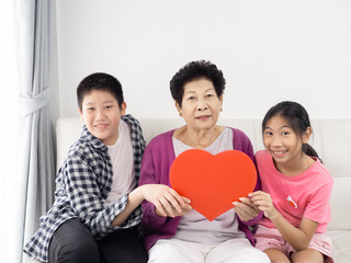 Happy Asian senior woman holding red heart shape sign with her grandchildren at home, lifestyle concept.