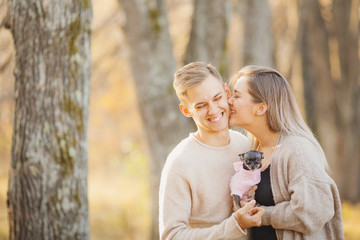 Autumn photo, couple in love hugs and holds dwarf dog on background of yellow leaves, girl bites man flirts cheek. Concept Childfree