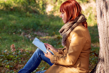 woman sitting under a tree in autumn park with tablet.