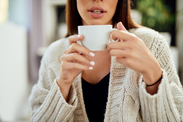 Caucasian woman dressed in beige sweater sitting on bed and holding fresh morning coffee.