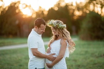 Young beautiful couple expecting a baby. Young beautiful pregnant woman with a wreath on her head holds on to a belly. Motherhood. Warm autumn.