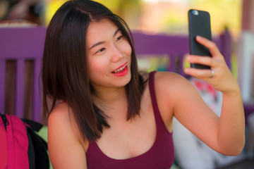 natural lifestyle take of young beautiful and happy Asian Japanese woman taking selfie portrait photo with hand phone smiling sitting outdoors enjoying Summer holidays