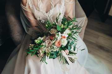 Bride is sitting on sofa, holding a bridal bouquet of roses wearing a wedding dress. Closeup