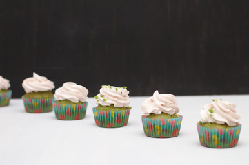Green cupcakes with cream hat in multi-colored papers on a white table on a dark background. Focus on cake, depth of field effect