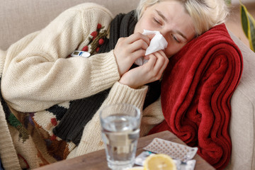 Girl in a sweater sneezes while sitting on a sofa. Colds and flu. The patient caught a cold,...