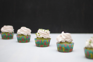 Green cupcakes with cream hat in multi-colored papers on a white table on a dark background. Focus on cake, depth of field effect