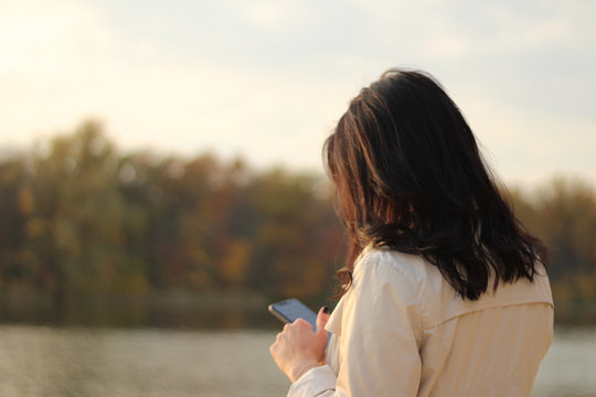 White dark haired girl looks at the cellphone and swipes a finger on the phone screen on a background of an autumn lake