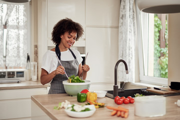 Attractive mixed race woman in apron mixing vegetables in bowl while standing in kitchen at home.