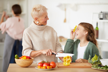 Little girl having fun while her grandmother cutting fresh pepper on cutting board they smiling to each other while standing in the kitchen