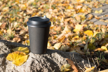 A black paper Cup of hot coffee stands on a stone, against a blurred background of autumn yellow leaves