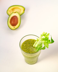 Green smoothie with a stalk of celery in a glass cup. On the table is an avocado.