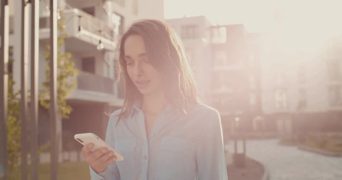 Caucasian happy and pretty young woman standing in the sunlights at the nice living block street and typing on the smartphone. Outdoors.