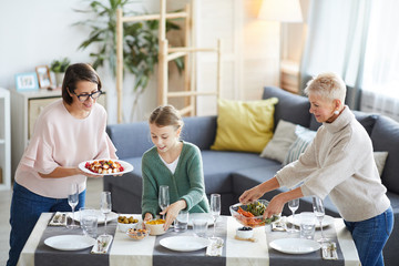Family of three putting different dishes on the dining table and they preparing for family dinner in the living room at home