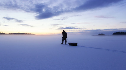 Man silhouette walk on the ice surface covored by snow at evening. Frozen Lake and forest...