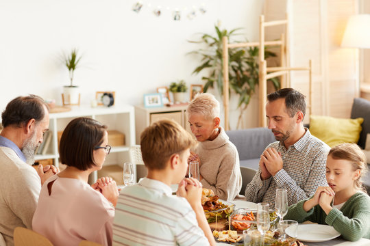 Big Family With Their Eyes Closed Praying Together Before Dinner At The Dining Table In The Living Room At Home